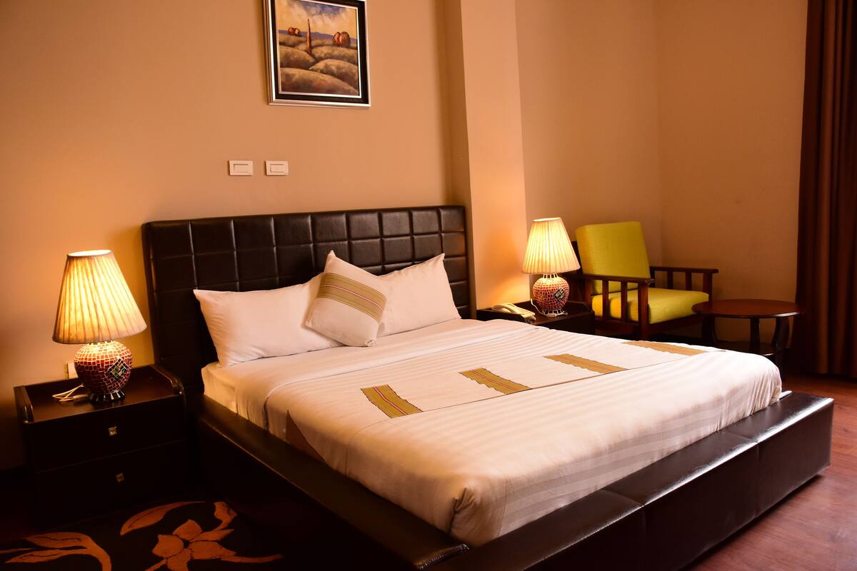 A hotel room with a large bed, two bedside lamps, a chair, a small table, and a painting on the wall.