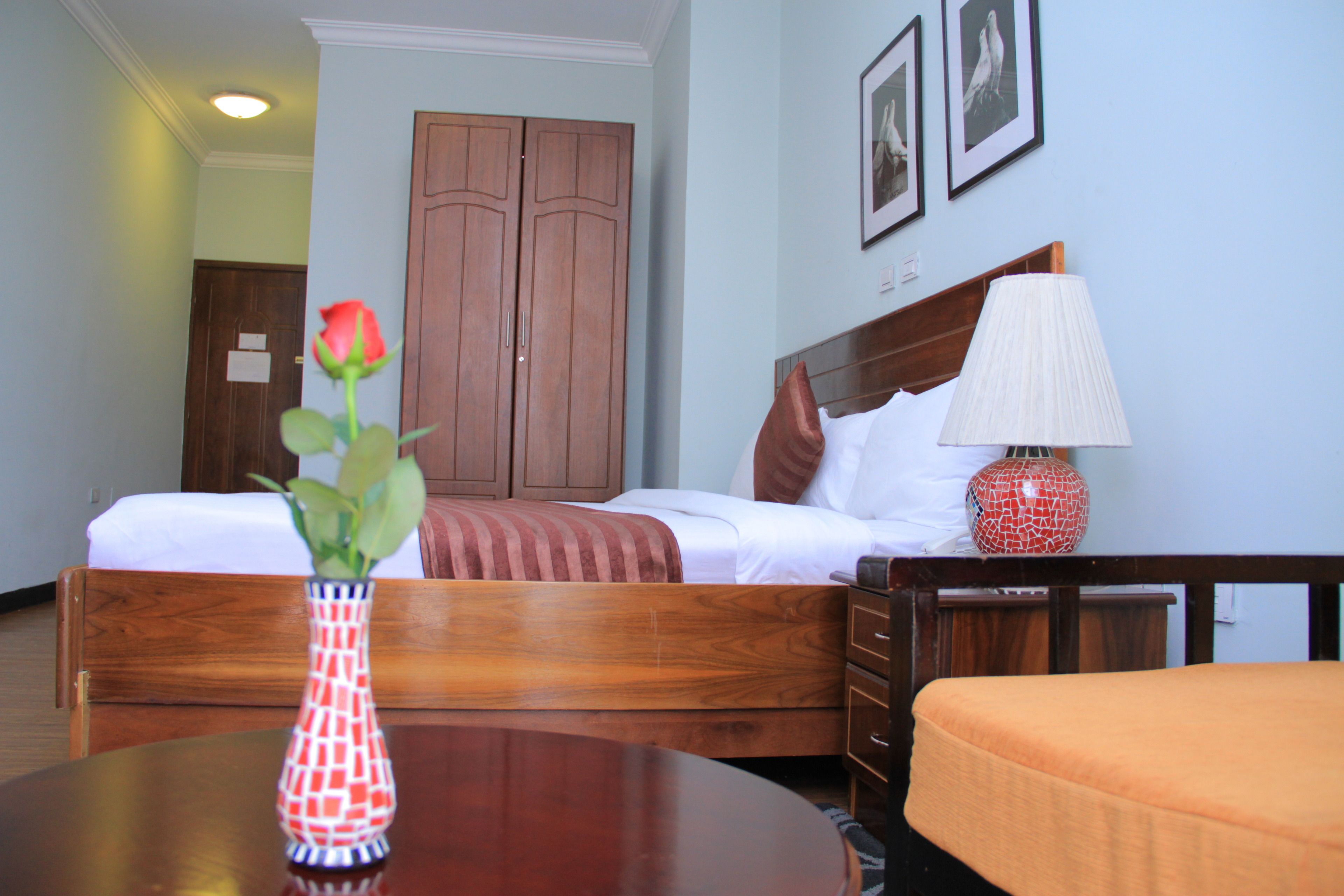 A hotel room with a wooden bed, a bedside table with a lamp, a vase with a rose, and a wooden nightstand.