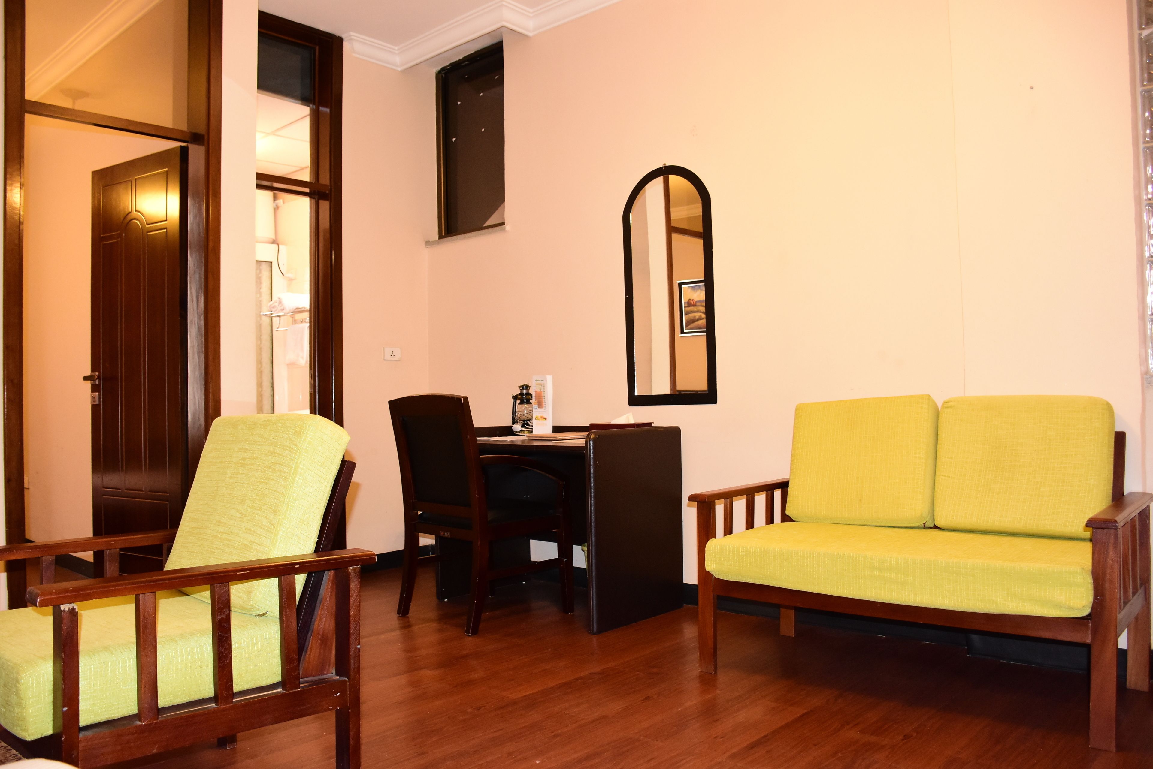 Une pièce avec un plancher en bois, un canapé jaune, une chaise en bois, un bureau avec une chaise et une porte.