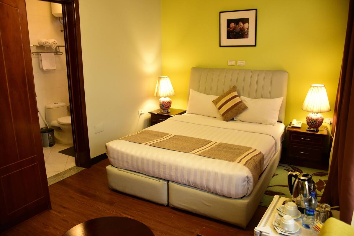 A hotel room with a bed, bedside tables, a nightstand, a framed picture on the wall, and a bathroom visible through an open door.