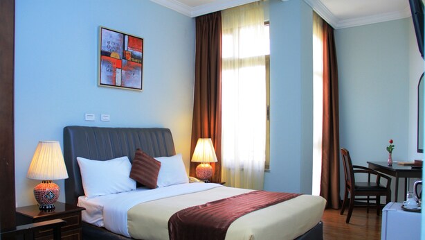 A hotel room with a bed, two bedside lamps, a desk with a chair, a window with curtains, and a painting on the wall. - Hotel Lobelia (Addis Ababa)