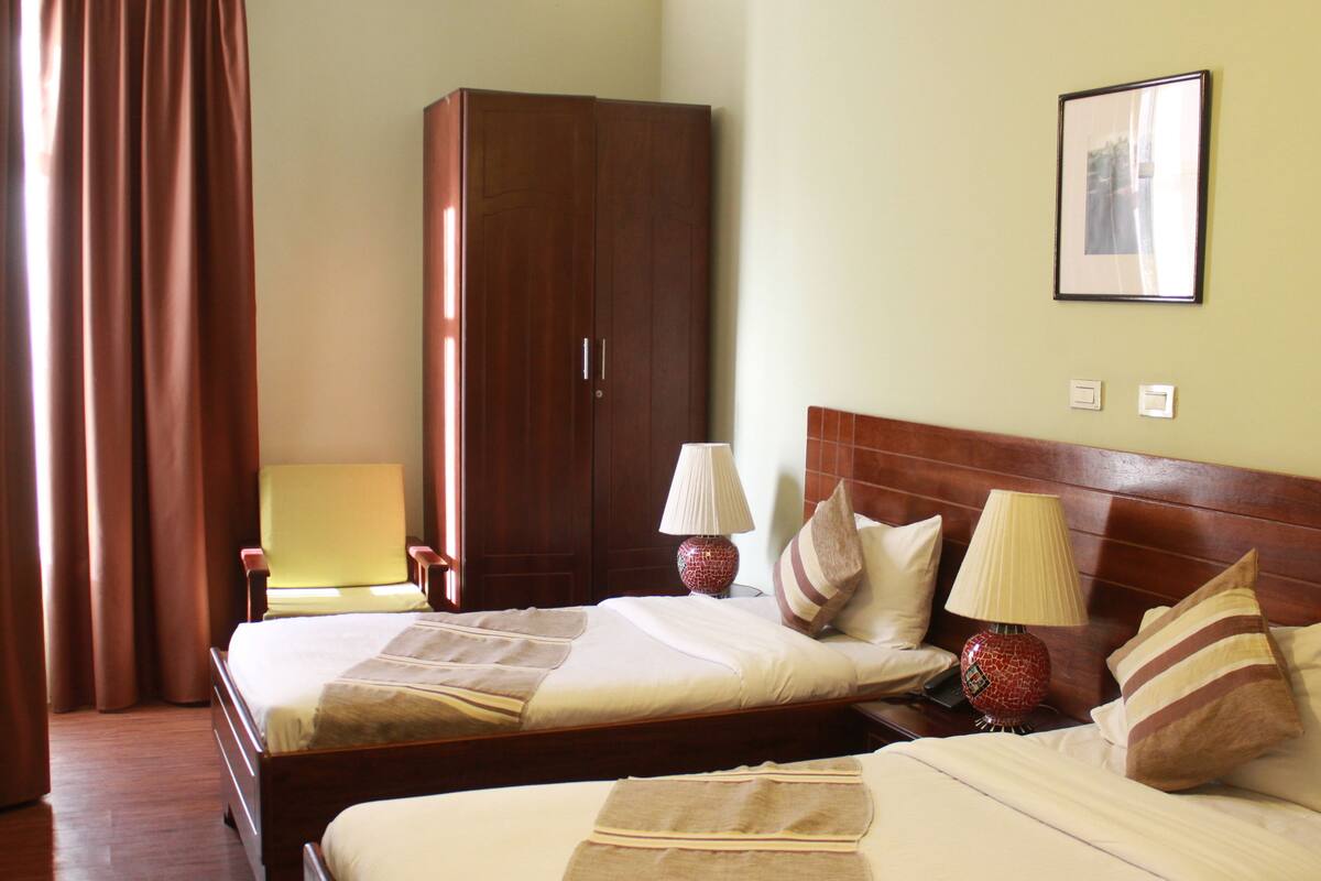 A hotel room with two beds, a wardrobe, a bedside table with lamps, and a framed picture on the wall.