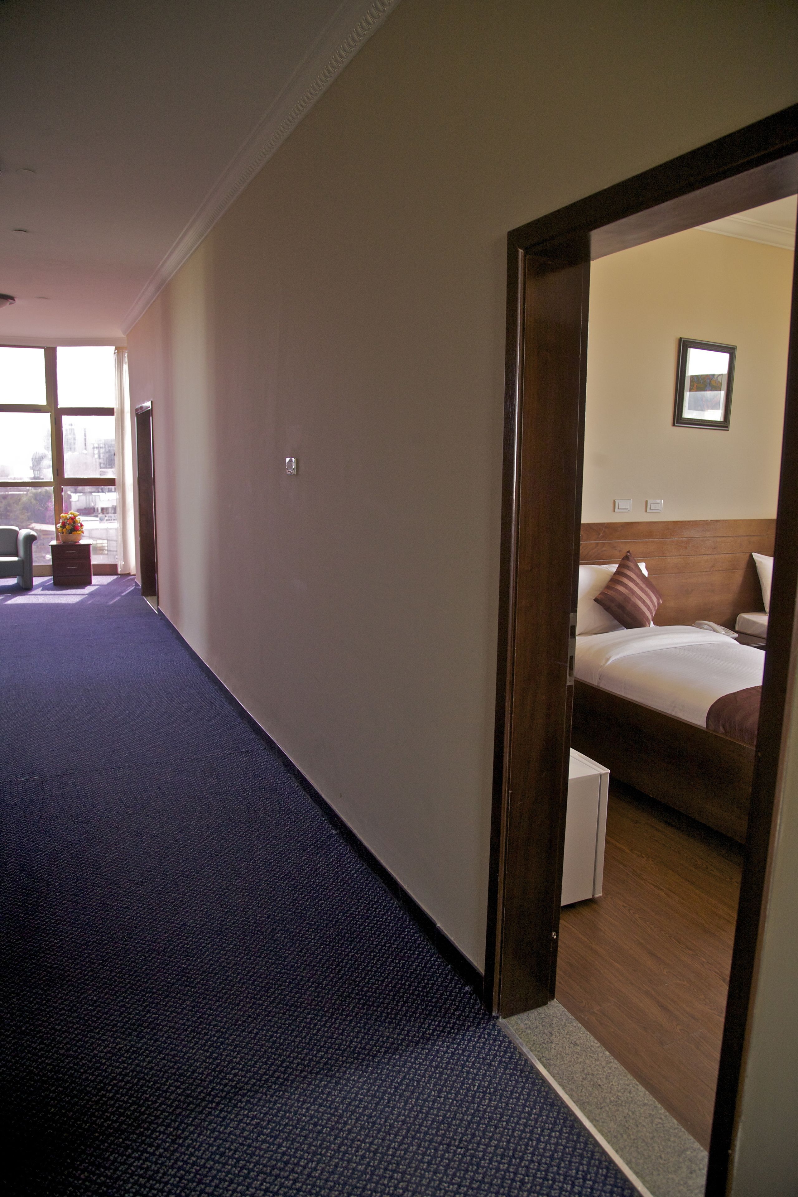 Un pasillo de hotel con suelo alfombrado, una cama con cabecera de madera y una mesita con un jarrón de flores.