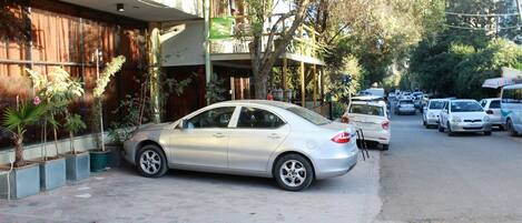A street view with parked cars, a building with a balcony, and potted plants.