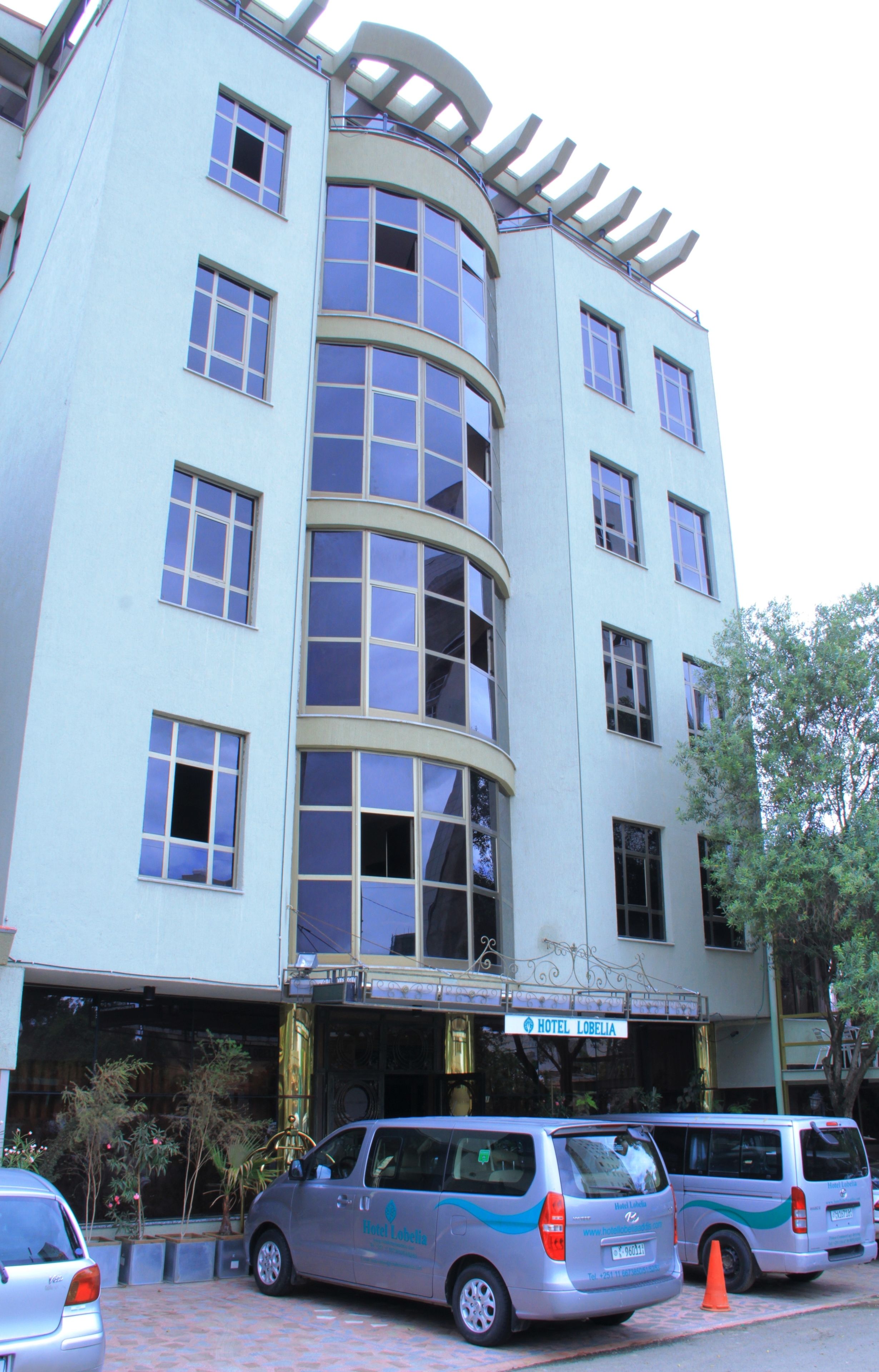 A multi-story hotel with a curved facade, large windows, and a sign indicating it is Hotel Lobelia. There are several parked vehicles in front.