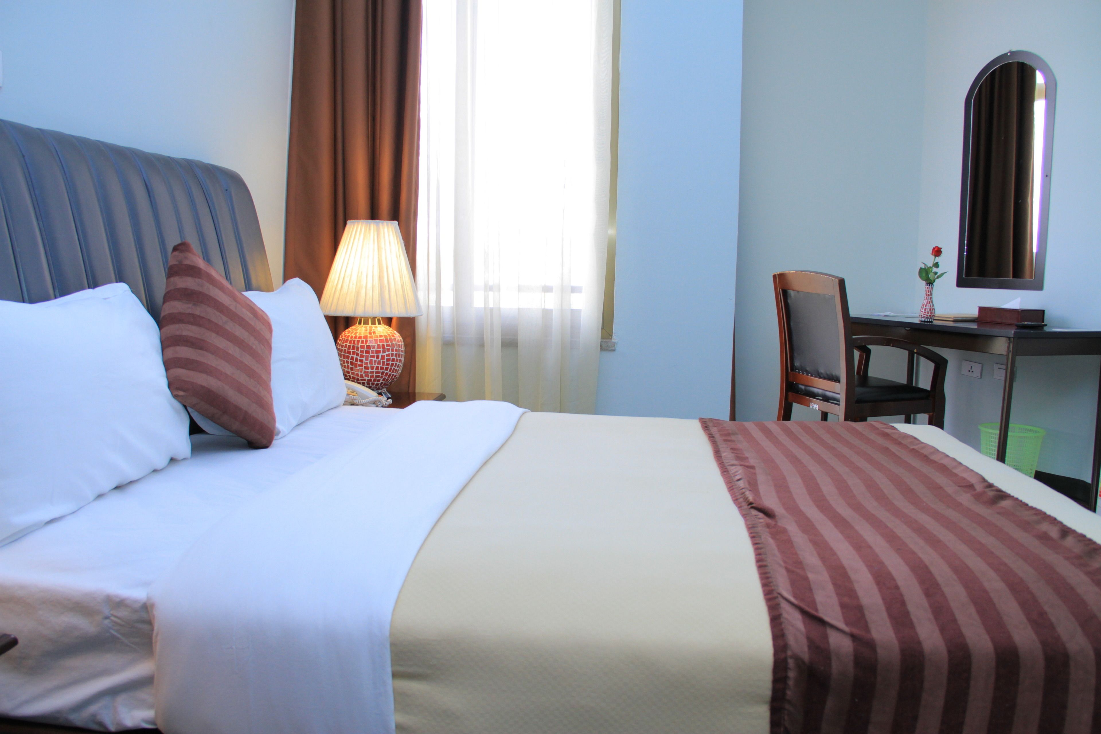 Una habitación de hotel con cama, escritorio, silla, lámpara y ventana con cortinas.