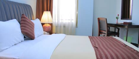 Una habitación de hotel con cama, escritorio, silla, lámpara y ventana con cortinas.
