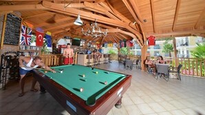 A pool table in a wooden-roofed room with flags, a bar area, and outdoor seating.