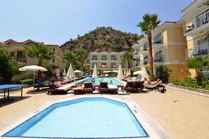 Zona de piscina con sillones de descanso y sombrillas, rodeada de edificios con balcones y una montaña al fondo.