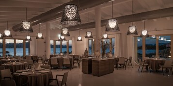 Comedor con mesas dispuestas para el servicio, amplias ventanas con vistas al agua y lámparas colgantes.