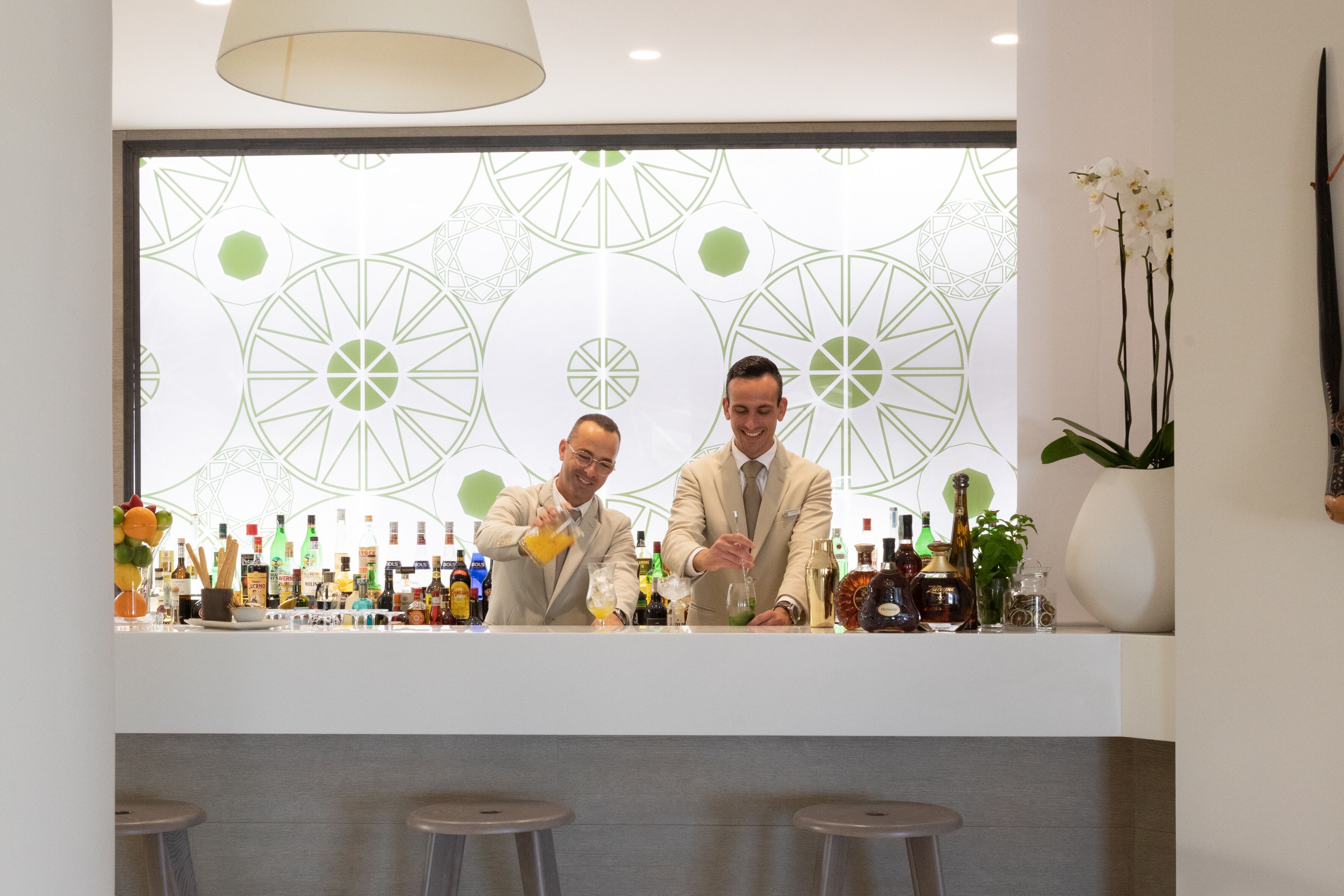 Zona de bar con dos bartenders preparando bebidas, una variedad de botellas y una pared decorativa con diseños geométricos.