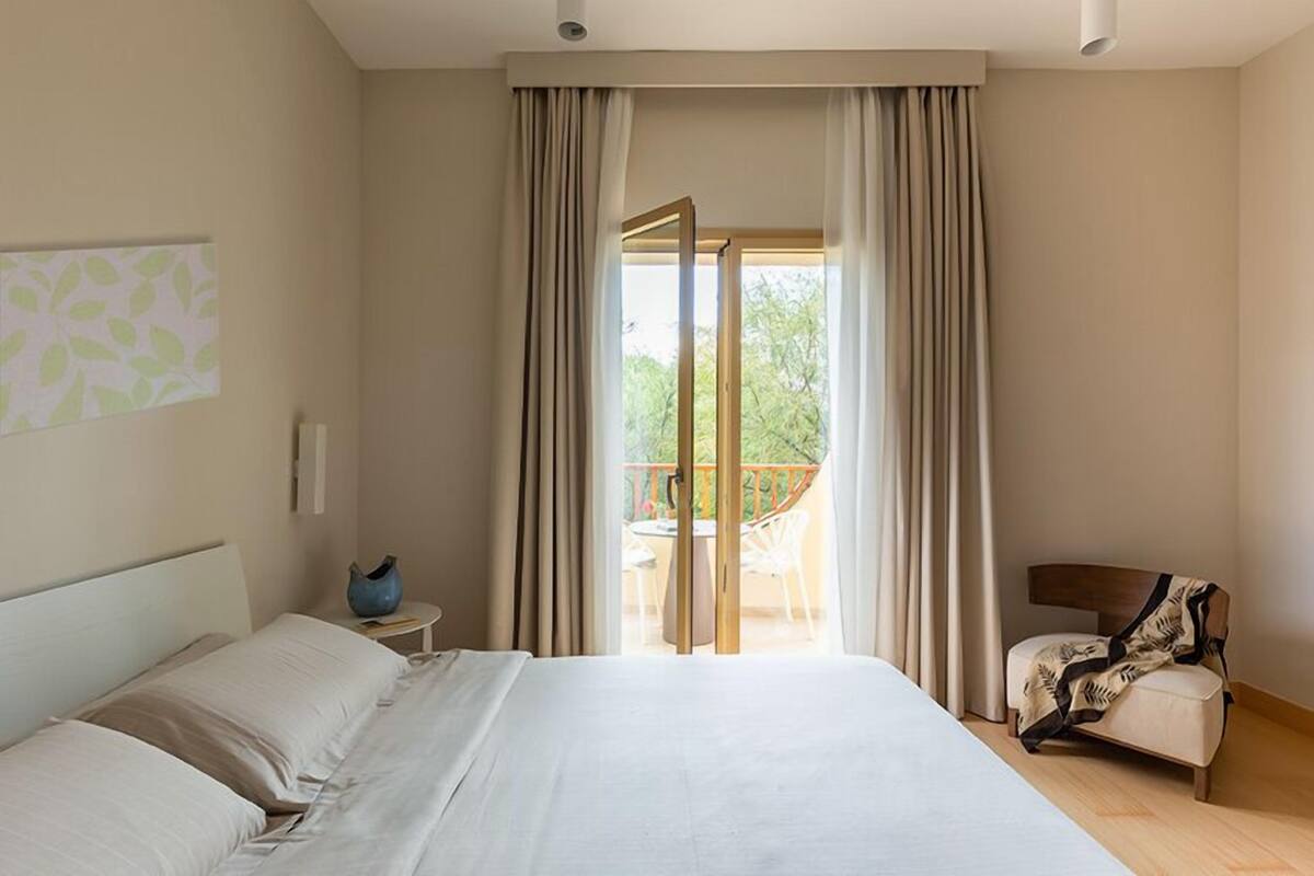 A hotel room with a large bed, a small table with a vase, a chair, and a balcony with a view of greenery.