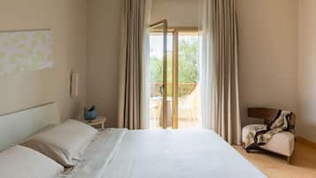 A hotel room with a large bed, a small table with a vase, a chair, and a balcony with a view of greenery.