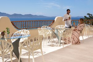 Una terrazza con sedie bianche e un tavolo in vetro, con vista sulla costa e montagne sullo sfondo.