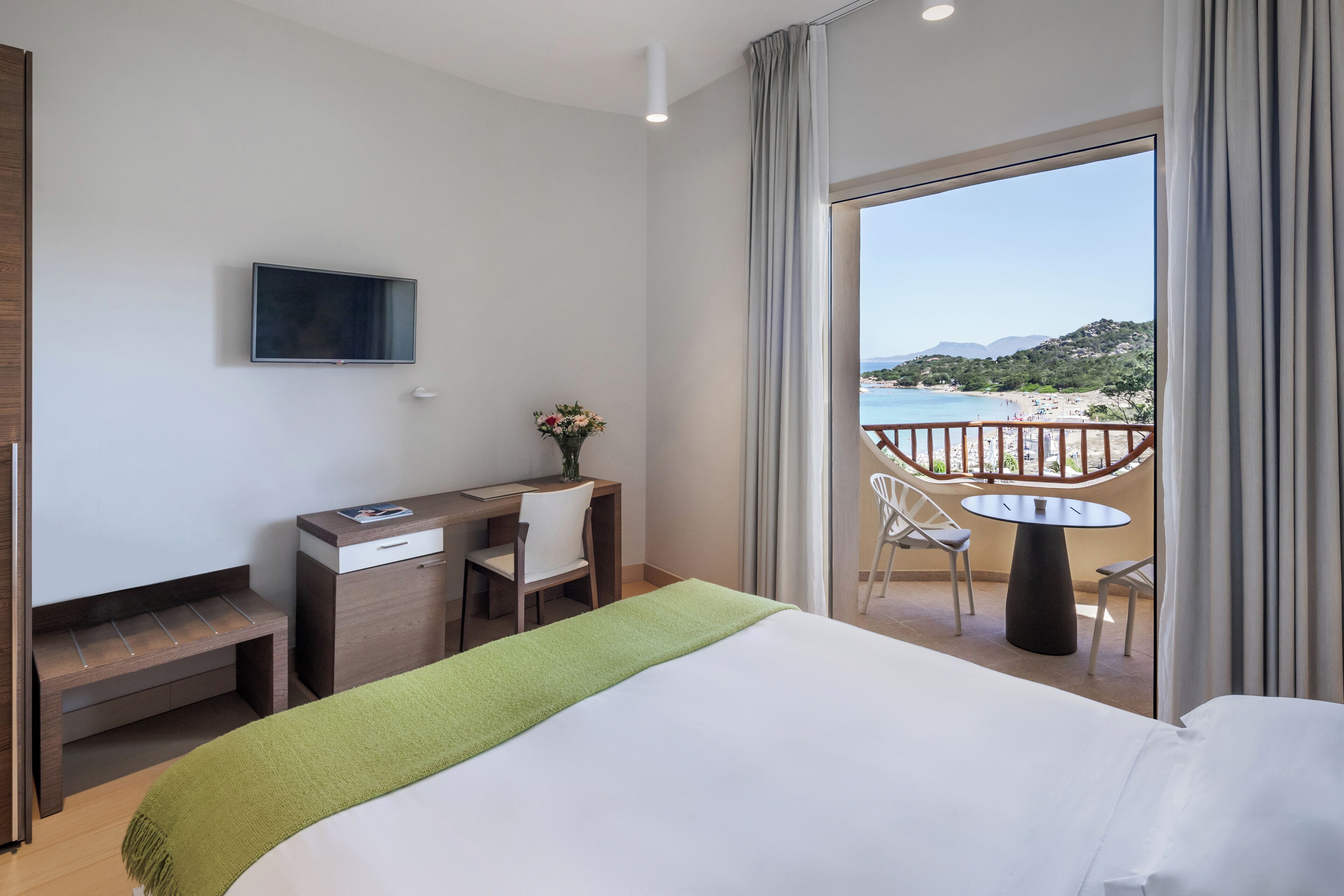 Een hotelkamer met een bed, een bureau, een stoel, een televisie en een balkon met uitzicht op het strand.