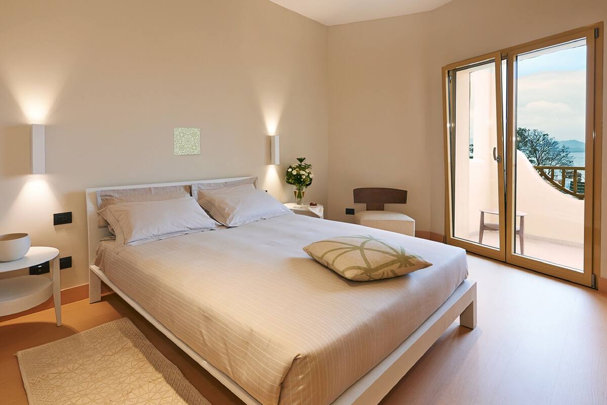 A bedroom with a bed, bedside tables, a nightstand, a vase with flowers, and a view of the outdoors through a sliding glass door.