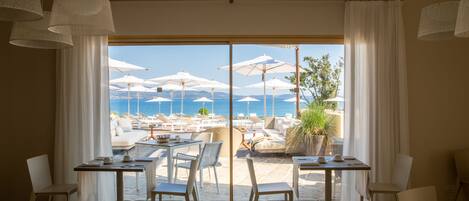 A room with a large window overlooking a beach with umbrellas and lounge chairs. There are several tables and chairs arranged inside the room.