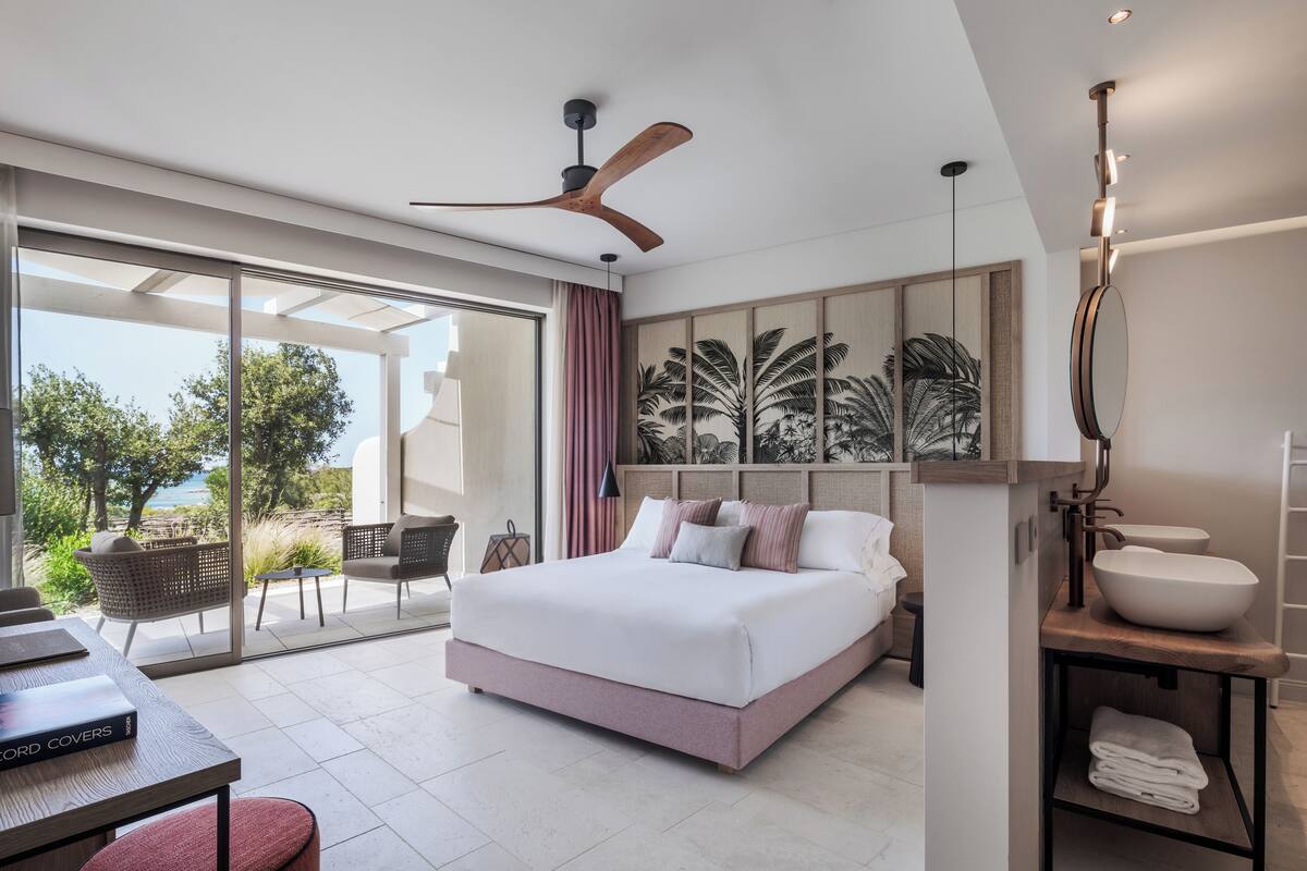 A modern bedroom with a large bed, a ceiling fan, a view of the outdoors, and a bathroom with a large sink and mirror.