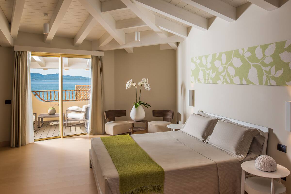 A bedroom with a bed, a green throw, a vase with flowers, a chair, a table, and a view of the sea through a balcony.