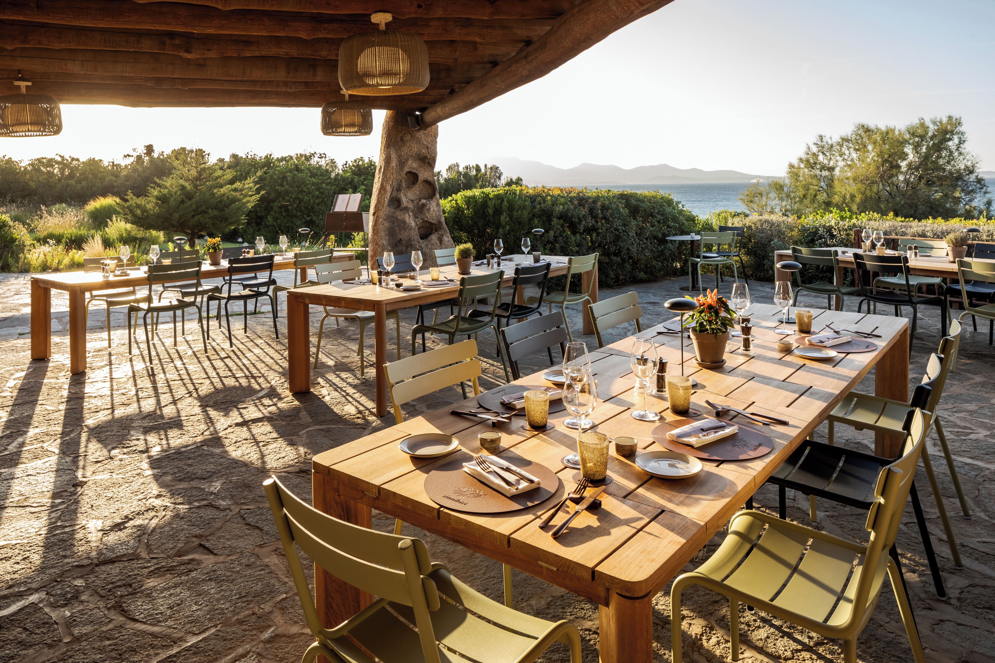 An outdoor dining area with multiple tables and chairs, set against a backdrop of greenery and a view of the sea.