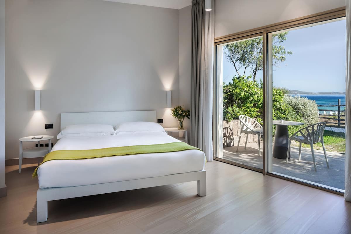 A modern bedroom with a large bed, bedside lamps, a nightstand, and a view of an outdoor patio with a table and chairs.
