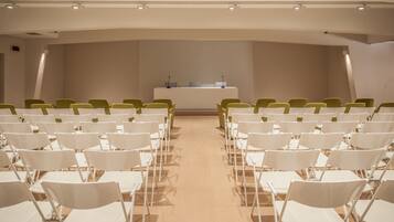 A conference room with rows of white chairs facing a long table with a laptop and microphone.