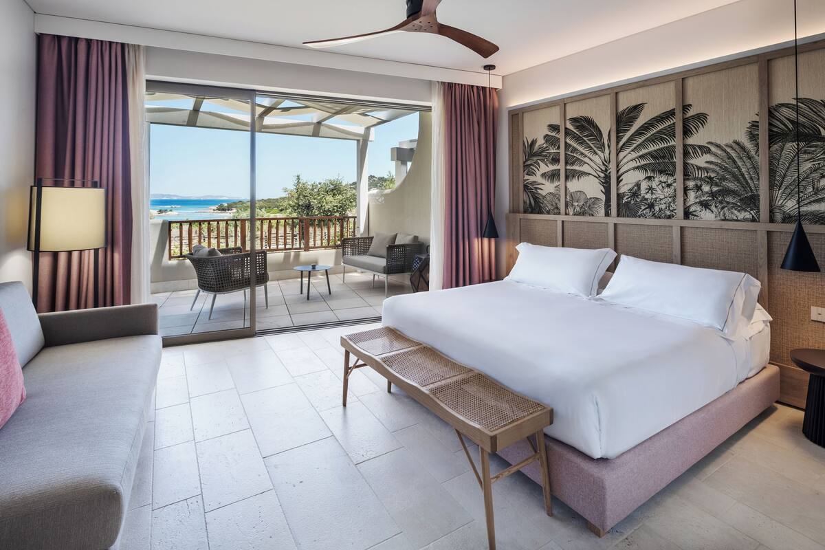 A hotel room with a large bed, a bench, a sofa, a small table, and a balcony with a view of the ocean.