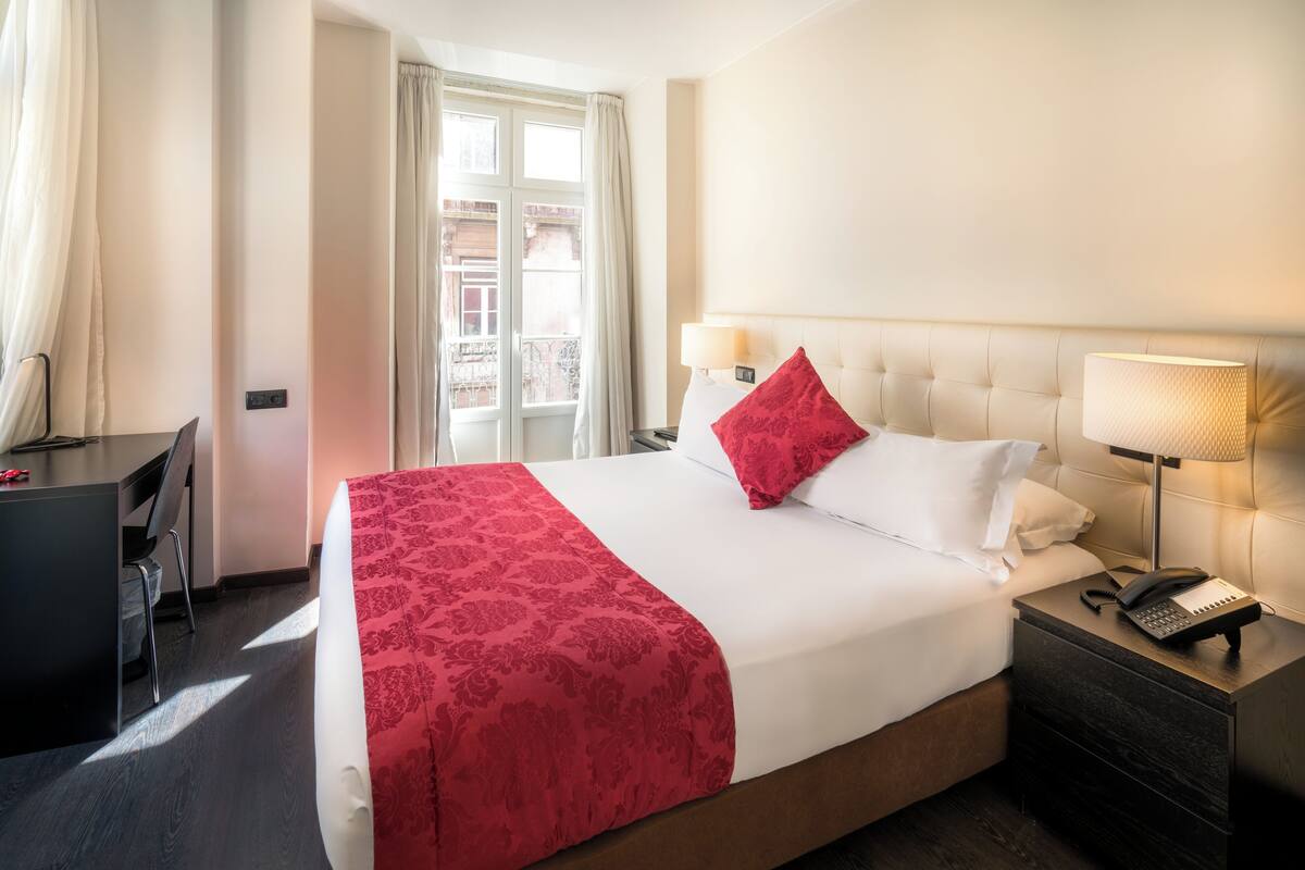 Een hotelkamer met een bed, nachtkastje met een lamp, een bureau en een raam met gordijnen.