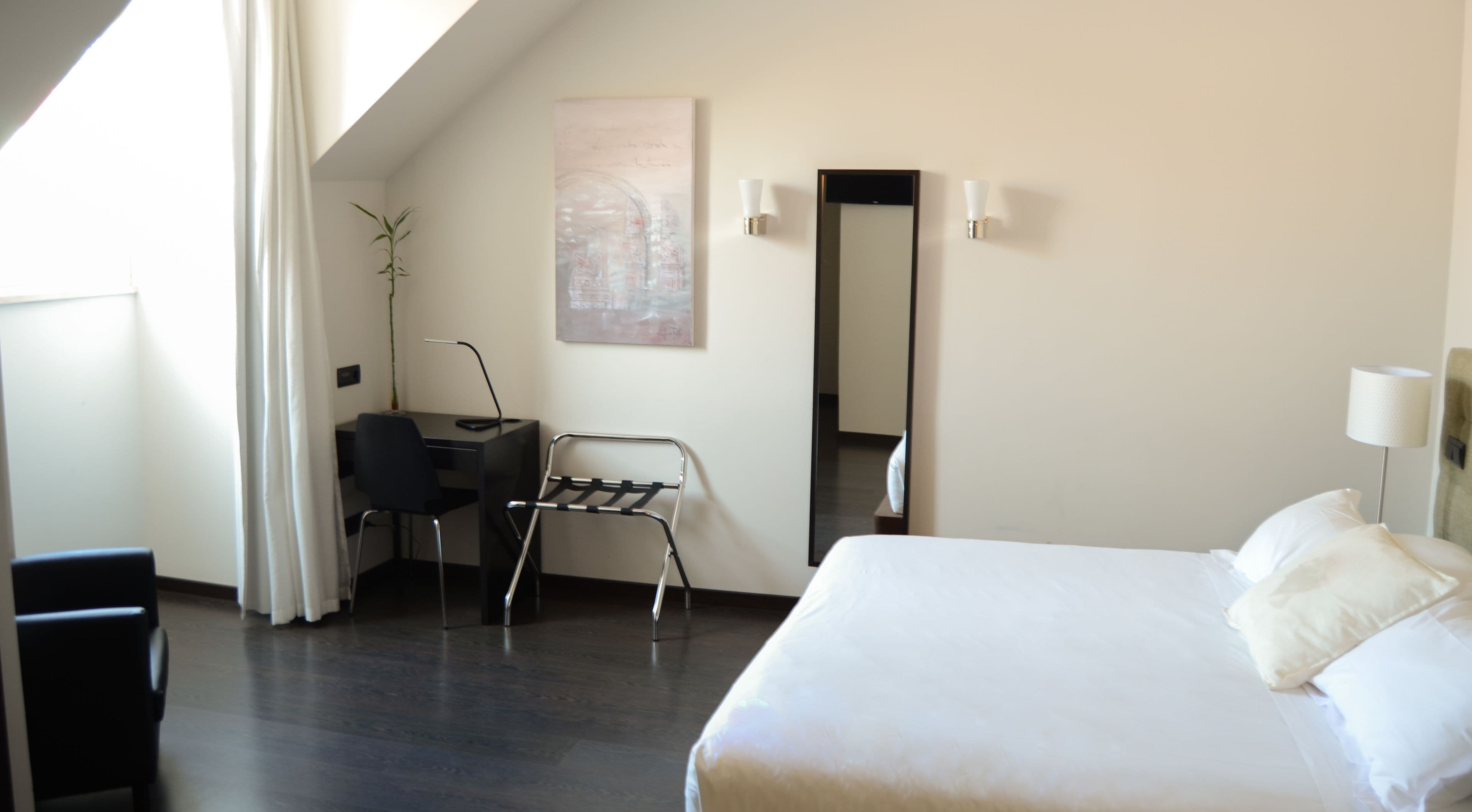 A modern hotel room with a large bed, a desk with a chair, a standing desk, a mirror, and a potted plant.