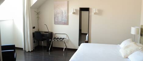 A modern hotel room with a large bed, a desk with a chair, a standing desk, a mirror, and a potted plant.
