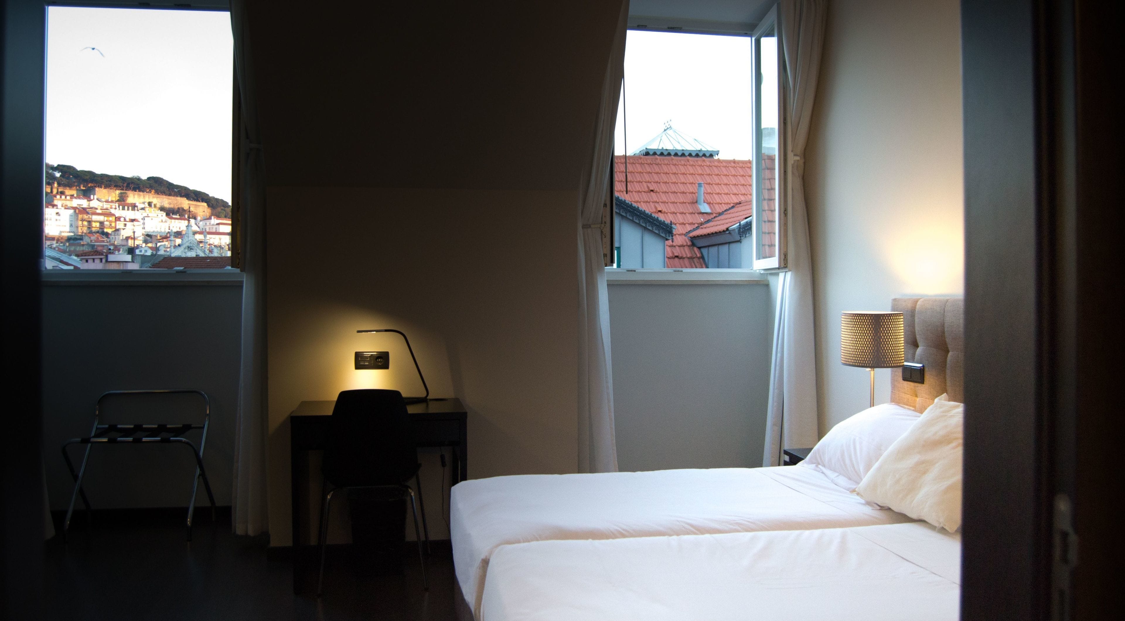 Habitación de hotel con cama, escritorio con lámpara, silla y ventana con vista a edificios.