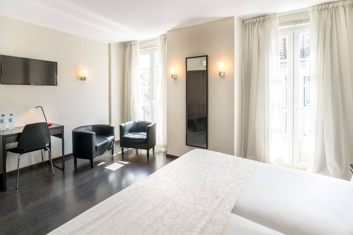Een hotelkamer met een bed, een bureau, twee stoelen, een televisie en een groot raam met gordijnen.