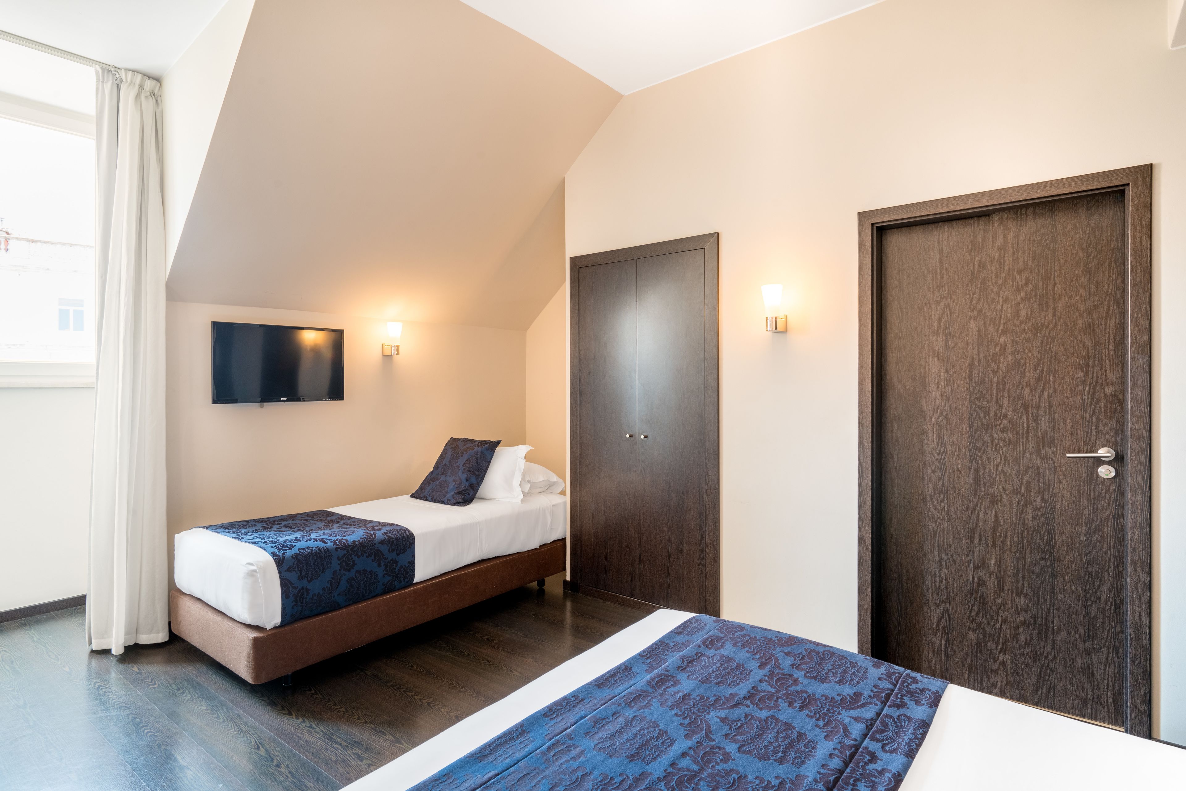 A bedroom with a bed, a wardrobe, a television mounted on the wall, and a window with curtains.