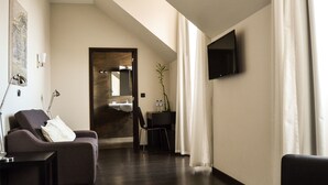 Une chambre d’hôtel avec un plancher en bois foncé, un canapé avec un coussin, un bureau avec une chaise, un téléviseur à écran plat fixé au mur et une porte donnant sur une salle de bain.