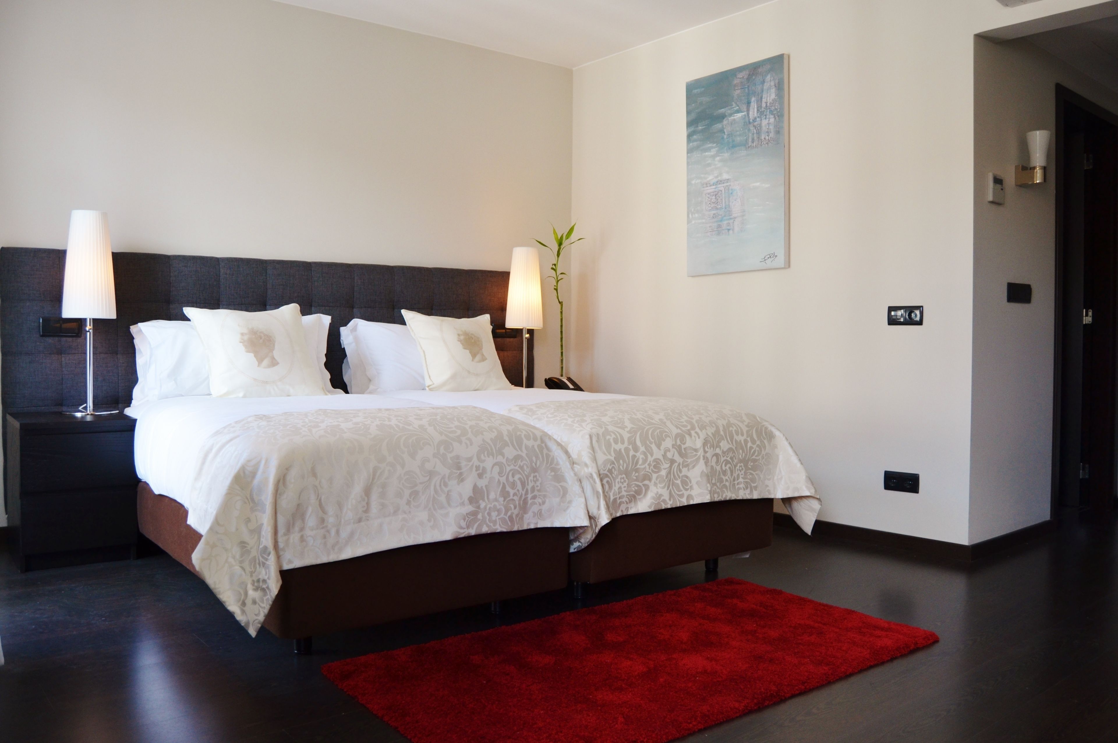 A modern hotel room with a large bed, two bedside lamps, a red rug, and a painting on the wall.