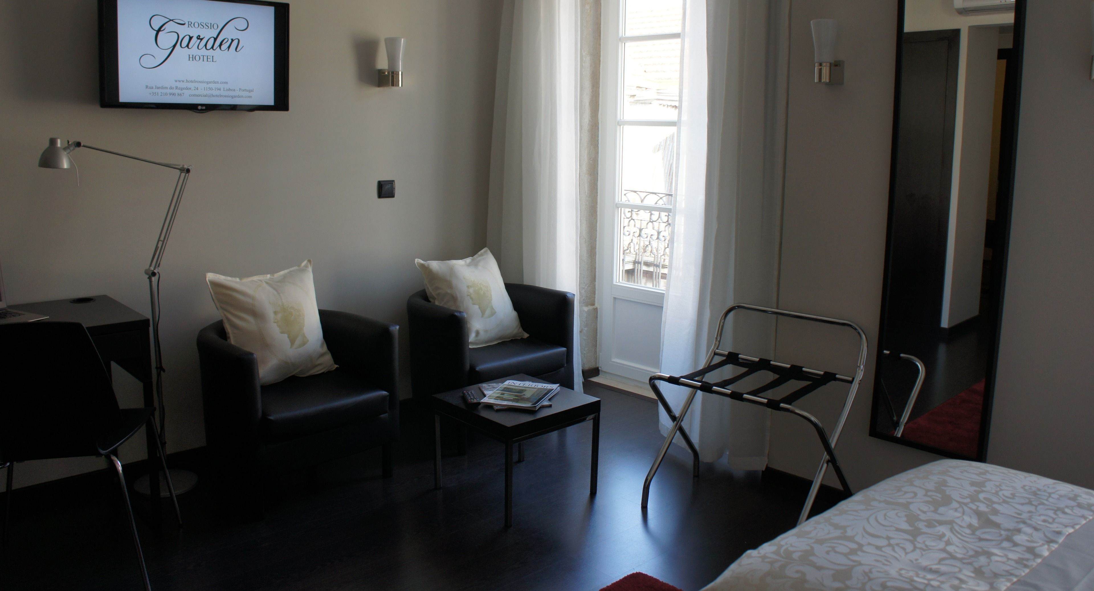 A hotel room with a bed, two sofas with pillows, a small table with a magazine, a floor lamp, a window with a view, and a wall-mounted TV.