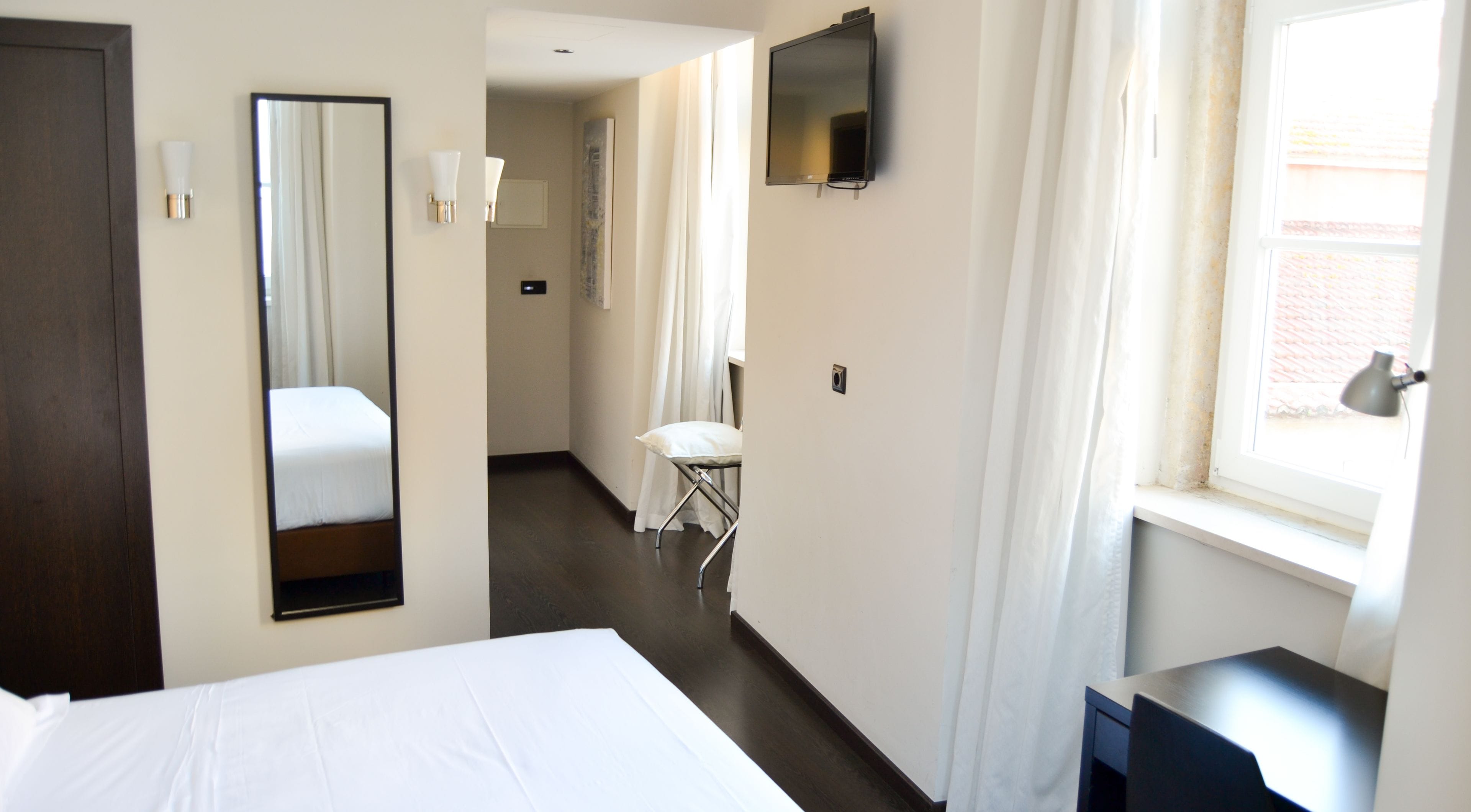 Une chambre d’hôtel avec un lit, une chaise, un bureau, un miroir, une télévision et une fenêtre avec des rideaux.
