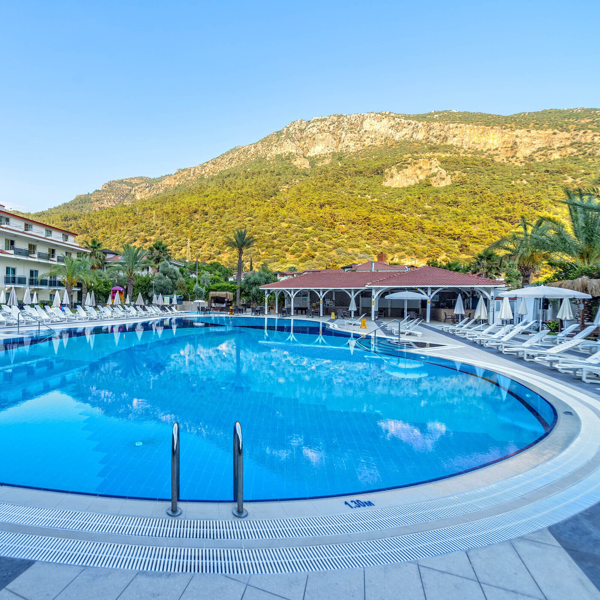 A swimming pool with lounge chairs and umbrellas, surrounded by buildings and a mountainous backdrop.