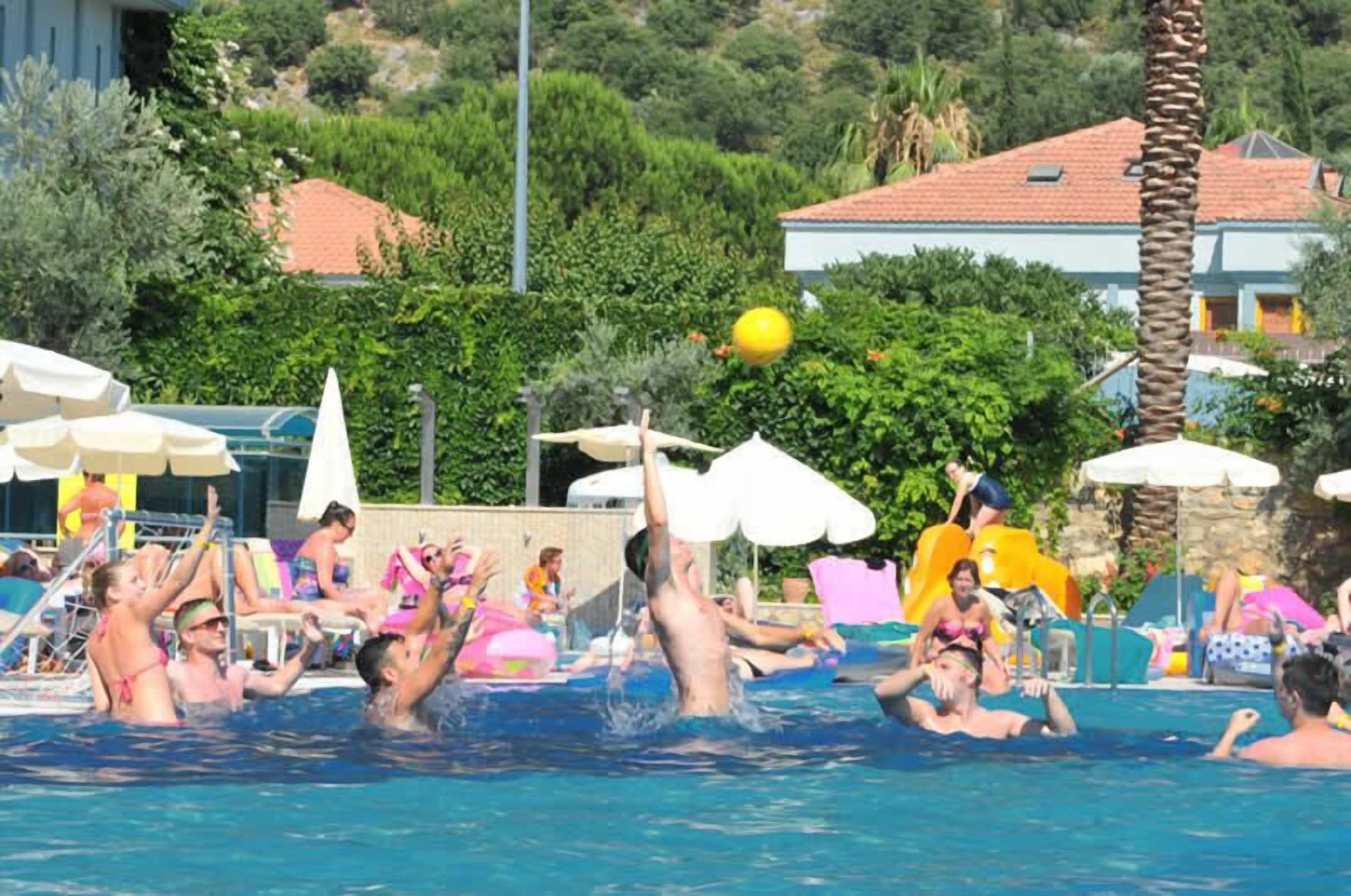 A swimming pool area with people playing, surrounded by umbrellas and greenery.