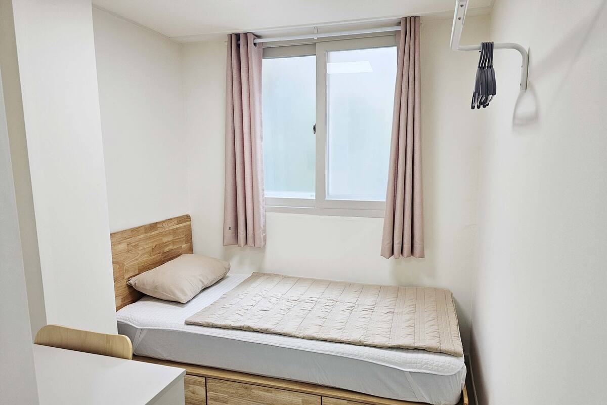 A small, neatly arranged bedroom with a single bed, a wooden headboard, a bedside table, and a window with curtains.