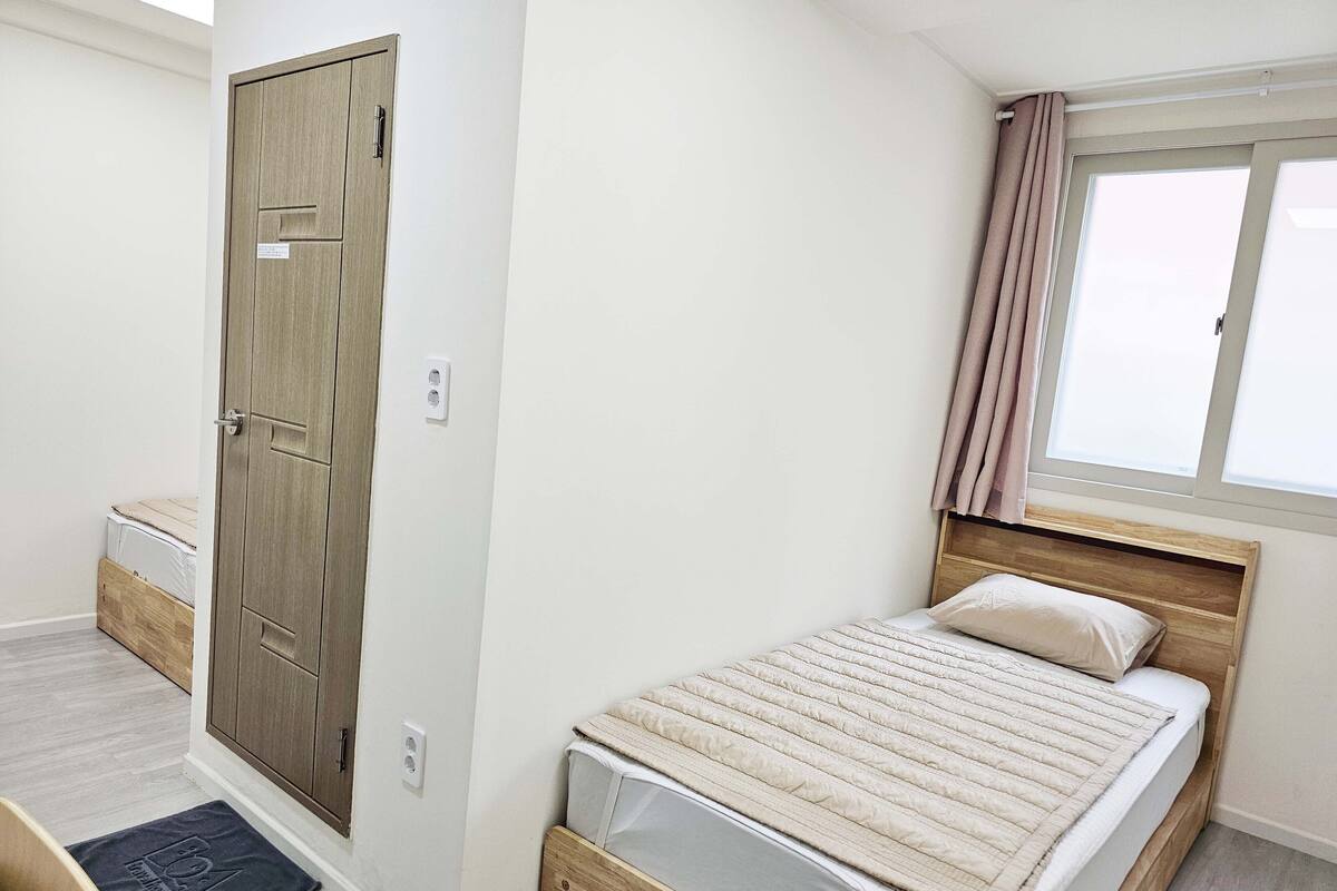 A bedroom with a wooden bed, a window with curtains, a door, and a small bedside table.