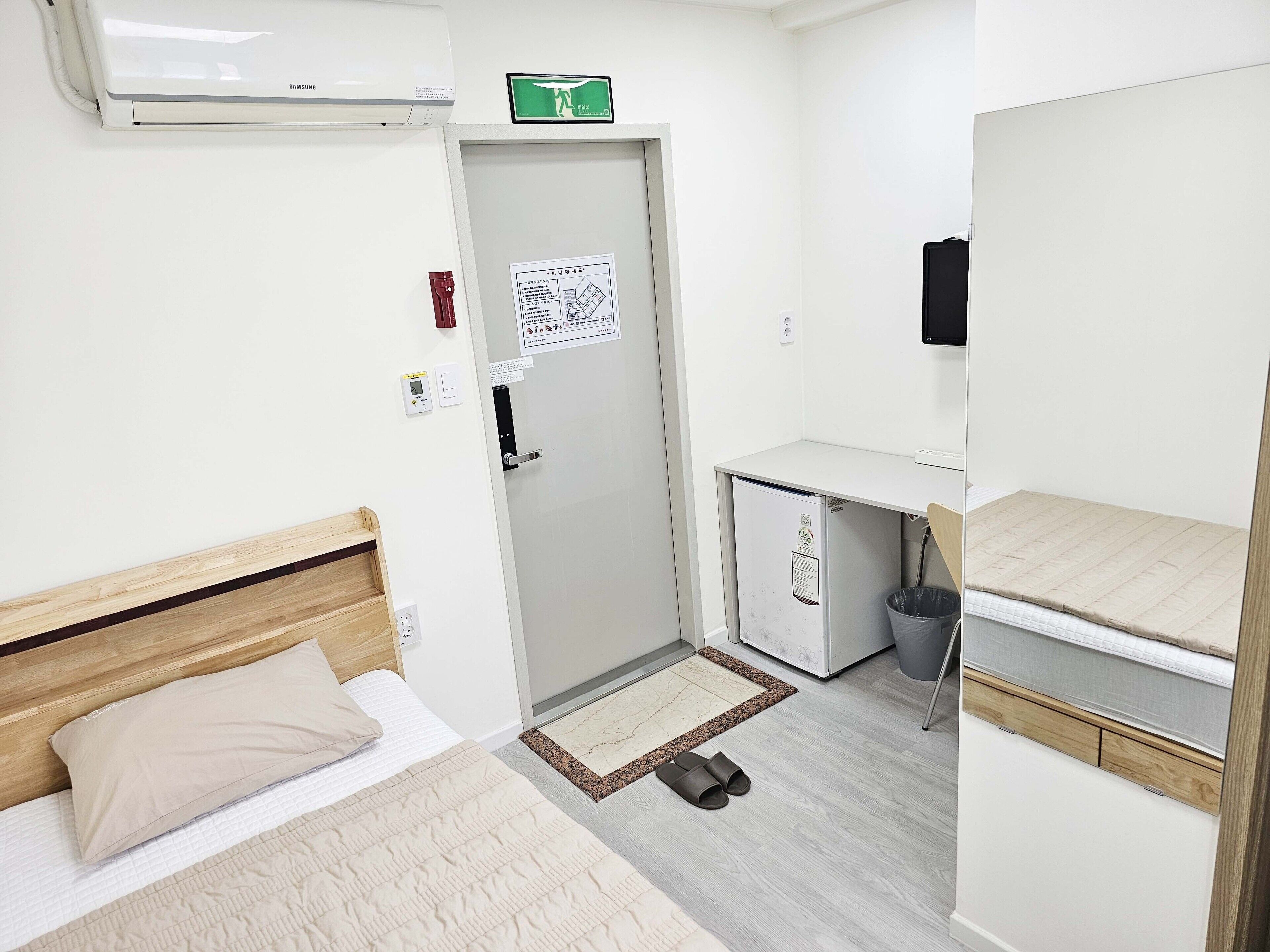 A compact hotel room with a bed, a desk, a small trash bin, and a mirror.