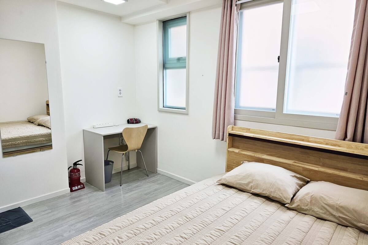 A bedroom with a bed, a desk, a chair, a window with curtains, and a fire extinguisher.