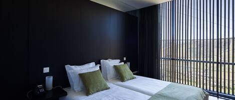 A modern hotel room with two beds, a nightstand, a lamp, and a window with vertical blinds.