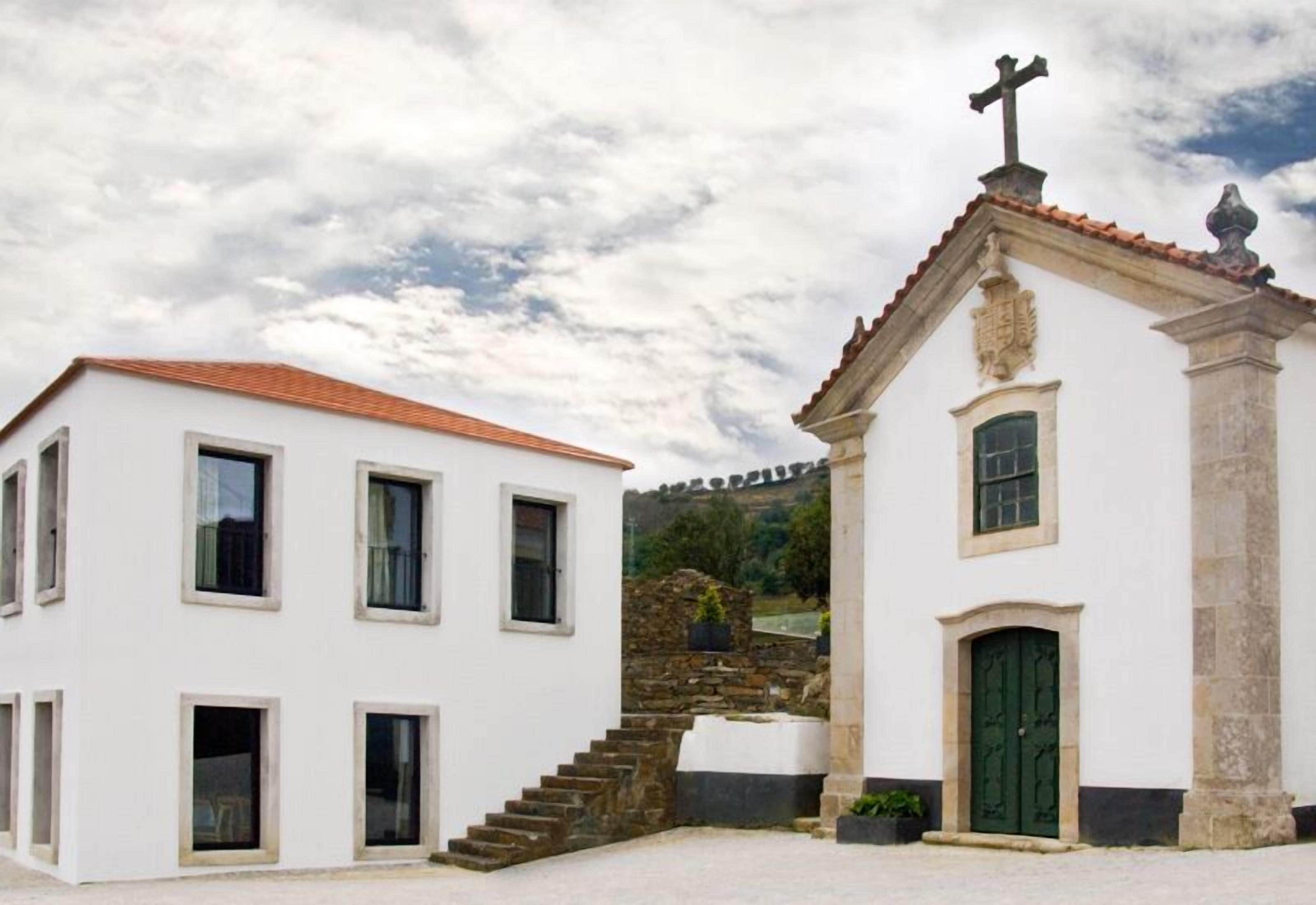 Un edificio blanco con techo de tejas rojas y una pequeña capilla con puerta verde y una cruz en la parte superior.