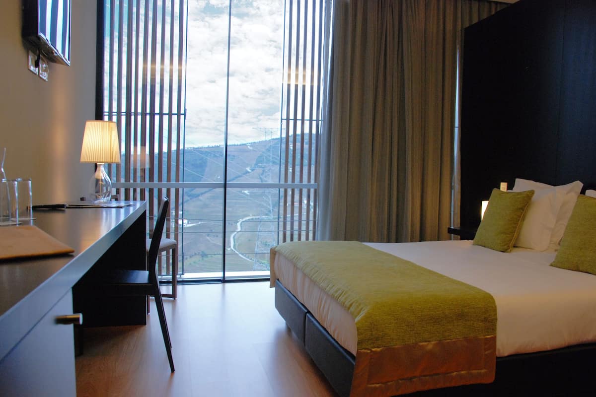 A hotel room with a bed, a desk, and a chair. There is a window with a view of a landscape.