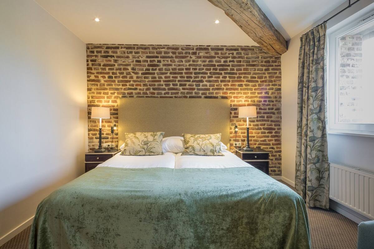 A hotel room with a brick wall, a large bed with a green bedspread, two bedside tables with lamps, and a window with curtains.
