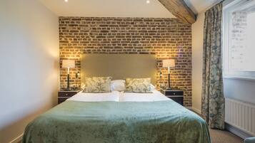 A hotel room with a brick wall, a large bed with a green bedspread, two bedside tables with lamps, and a window with curtains.