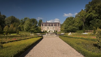 Un edificio grande e histórico con un jardín simétrico y un sendero de grava que conduce a la entrada.