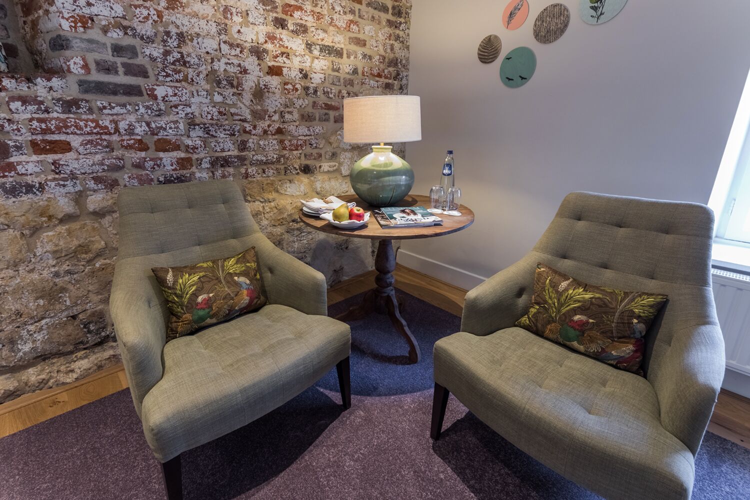 A room with two armchairs, a round table with a lamp, and a brick wall.
